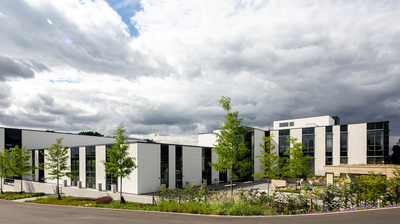 Moderne Bürogebäude mit weißer Fassade, großen Glasfronten und Bäumen im Vordergrund unter bewölktem Himmel