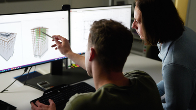 Person zeigt auf ein 3D-Gebäudemodell auf einem Monitor, digitale Bauplanung mit CAD-Software