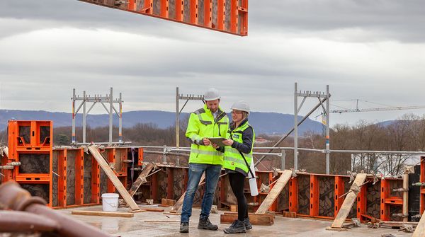 Architektin und Bauleiter in Warnschutzjacken besprechen einen Plan auf einer Baustelle mit Stahlträgern und Schalungen, im Hintergrund schwebt ein Metallträger am Kran.
