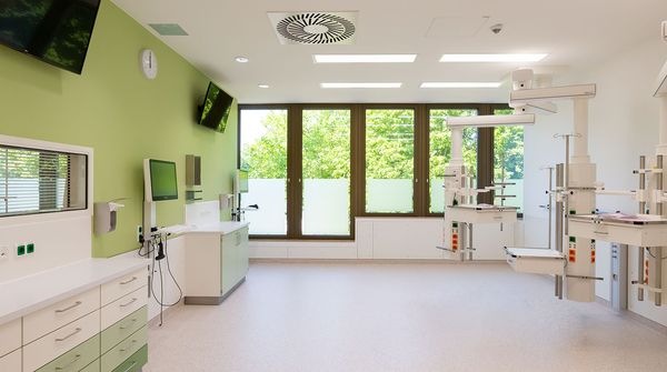 Innenansicht eines hellen Klinikraums mit grüner Wand, großen Fenstern und moderner medizinischer Ausstattung