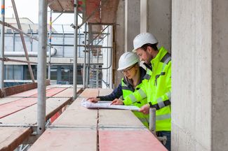 2 Architekten schauen sich gemeinsam auf dem Baugerüst die Baupläne an.